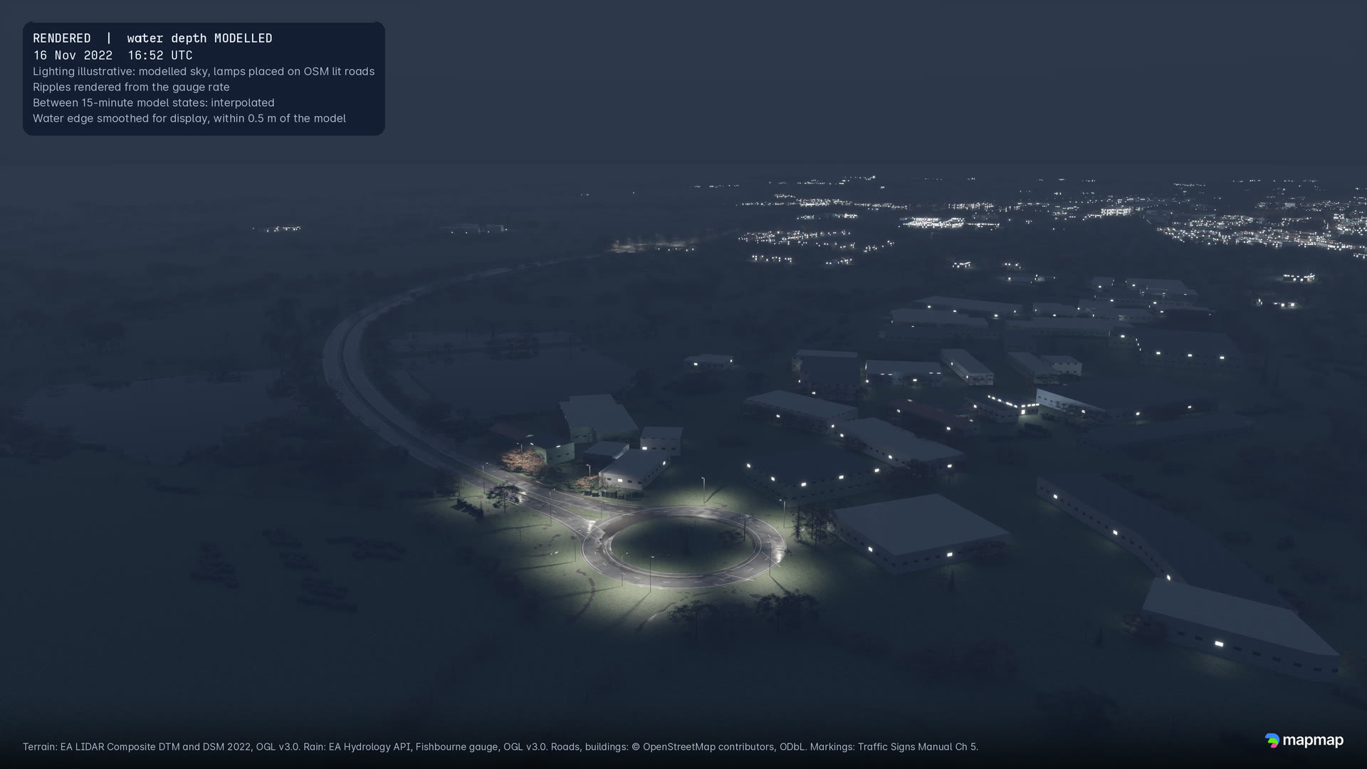 The bypass at dusk, 16:52 UTC, from high above: the lit roundabout, low buildings beside it and the lights of Chichester beyond.